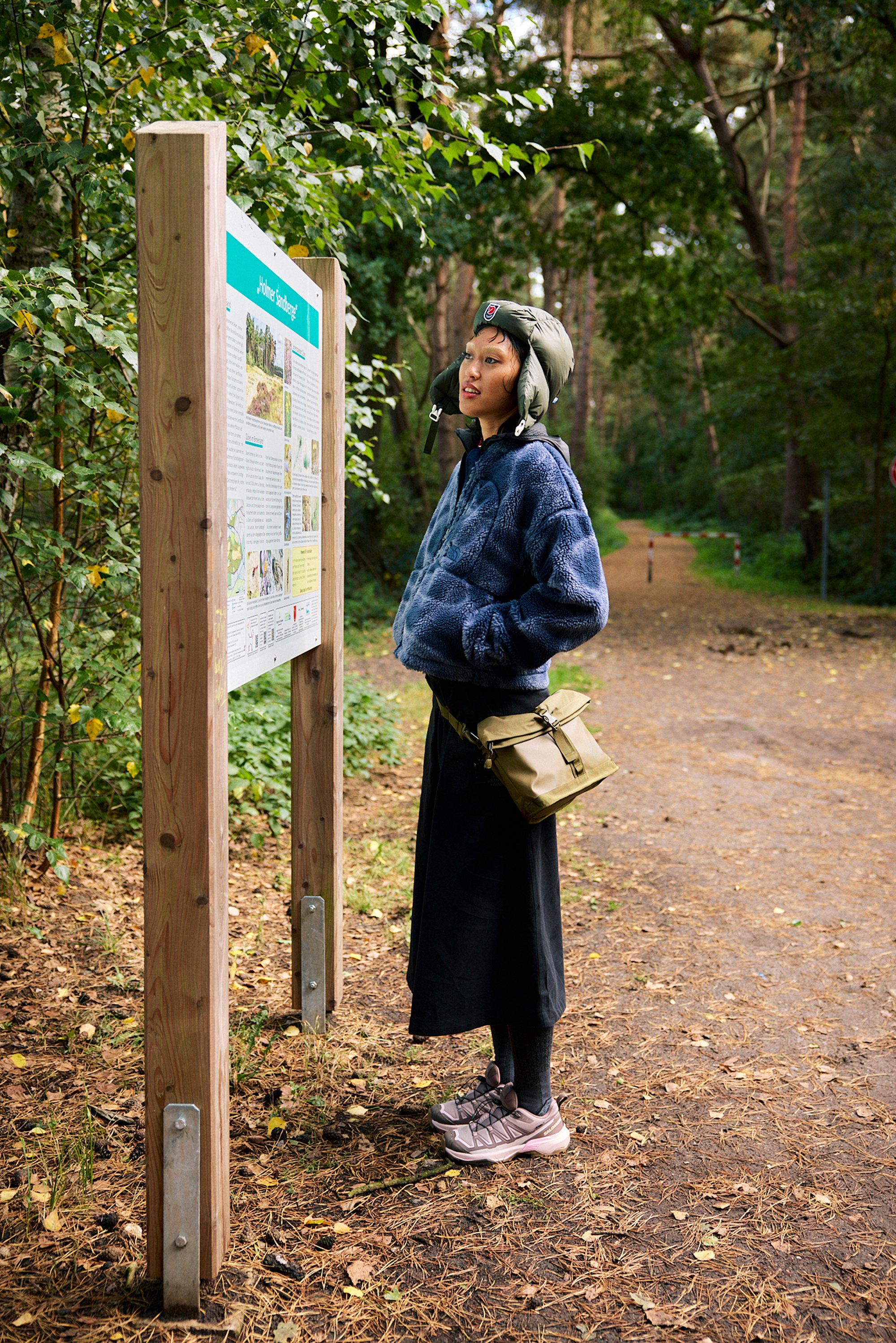 Blue fleece jacket, black skirt, and grey sneakers. Green helmet, and a beige crossbody bag. Wooden sign with informational graphics.