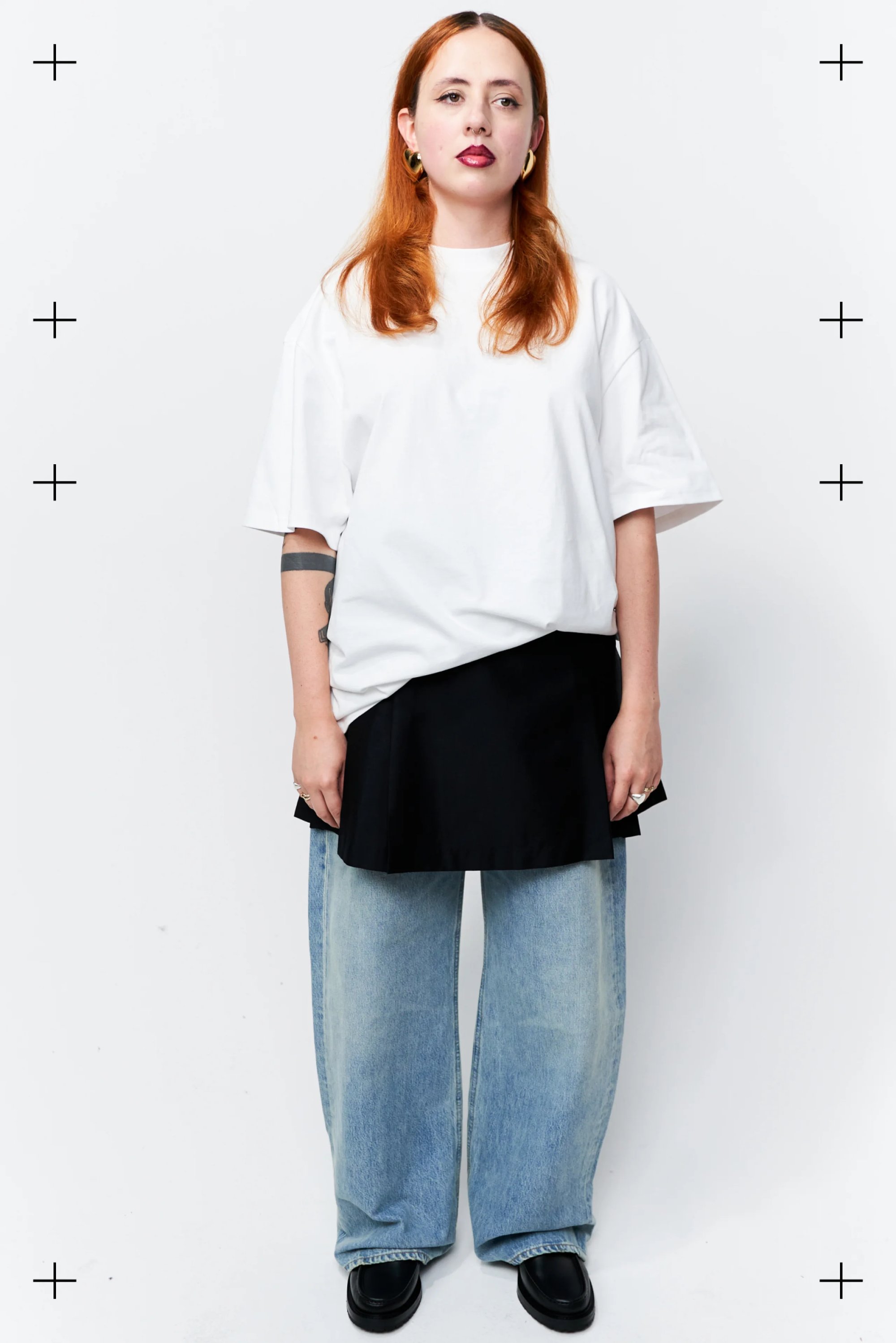 Person with long red hair stands in a white oversized t-shirt, black skirt, and light denim jeans, set against a plain white background.