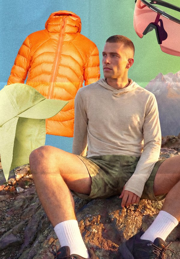 Mand i beige hættetrøje og grønne shorts sidder på klippefyldt jord, med orange dunjakke, grøn solhat og pink solbriller i nærheden.