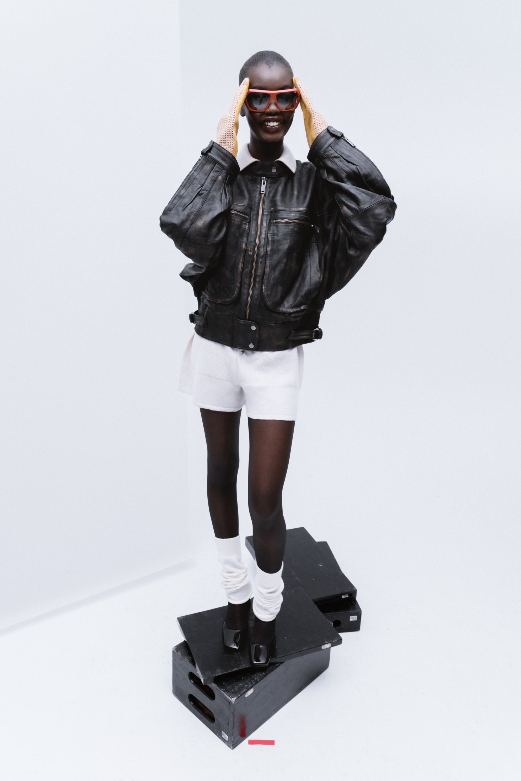Una modelo sonriente lleva una chaqueta de cuero negra, gafas de sol rojas y pantalones cortos blancos mientras está de pie sobre cajas negras contra un fondo claro.