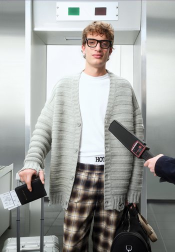 Cardigan gri deschis cu dungi și nasturi peste o cămașă albă, pantaloni în carouri, ținând un pașaport și o valiză, într-un cadru de securitate aeroportuară.