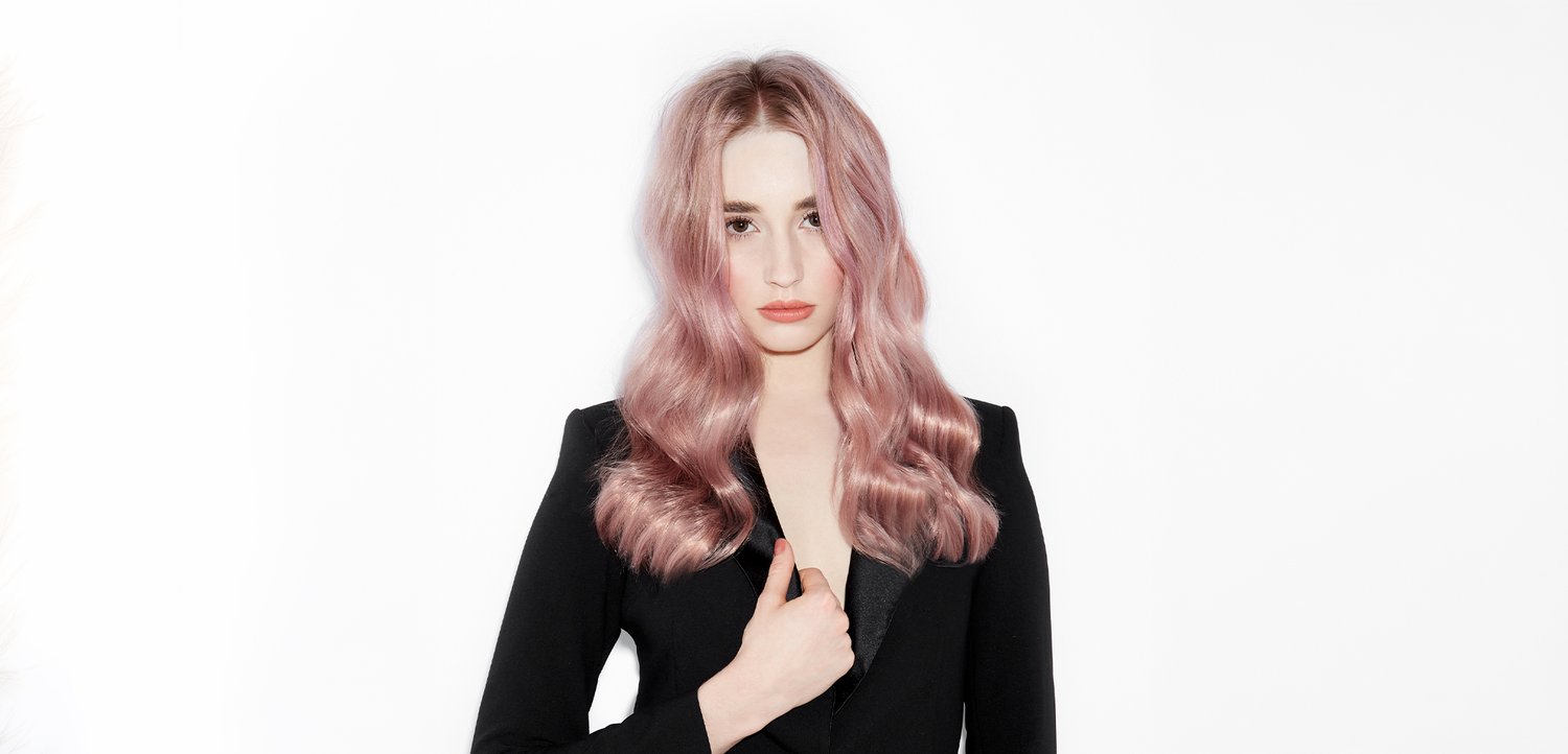 Junge Frau mit langen, welligen rosa Haaren, die einen schwarzen Blazer vor einem schlichten weißen Hintergrund trägt und direkt geradeaus schaut.