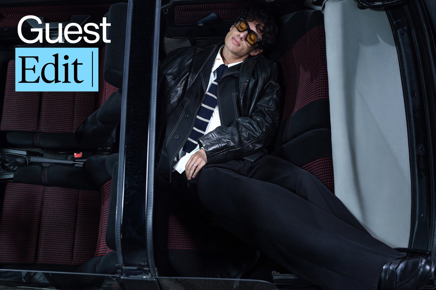 Una persona se relaja en un coche, vistiendo una chaqueta de cuero y gafas de sol, con una corbata a rayas con estilo. El interior del coche cuenta con asientos con acentos rojos.
