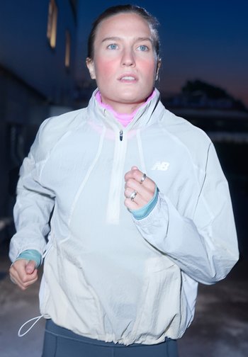 Γυναίκα με μπλε μάτια που τρέχει σε εξωτερικό χώρο κατά τη διάρκεια του λυκόφωτος, φορώντας ένα ανοιχτό γκρι μπουφάν New Balance και με ασημένιες βέρες στα δάχτυλά της.