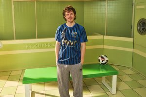 A man stands by a green bench in a brightly colored room, wearing a blue striped soccer jersey and gray pants, with a soccer ball nearby.