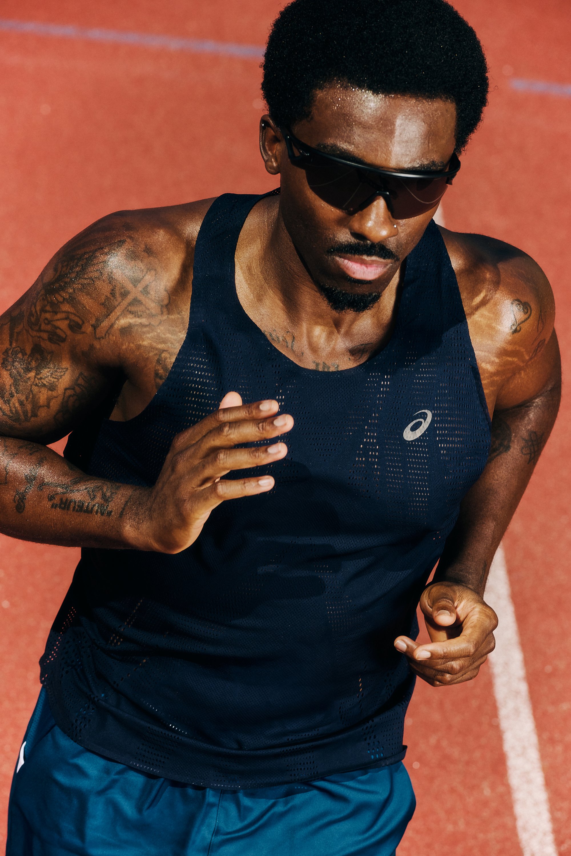 Man with tattoos runs on a red track, wearing sunglasses and a sleeveless navy shirt, showing determination and focus in his expression.