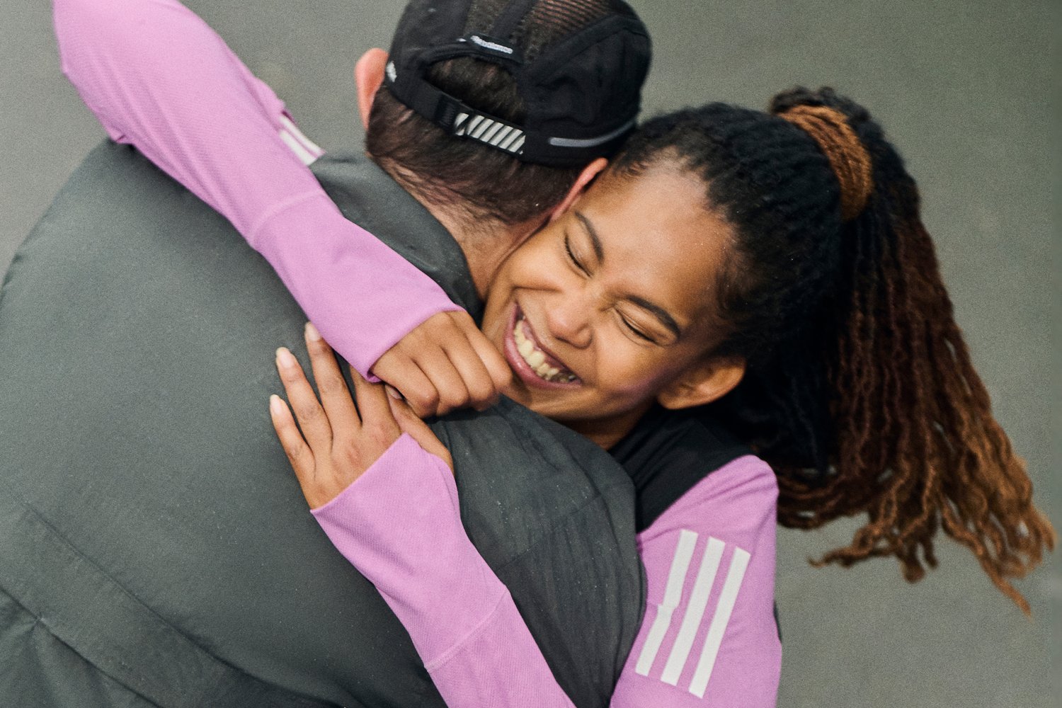 A woman with dreadlocks happily hugs a man from behind, both smiling. She wears a pink long-sleeve shirt, while he has on a grey jacket.