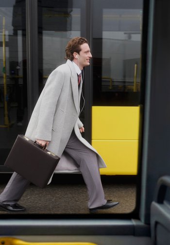 Grijze jas over een wit overhemd en een stropdas met patroon, grijze broek, zwarte schoenen, met een bruine aktetas, lopend langs een gele bus.