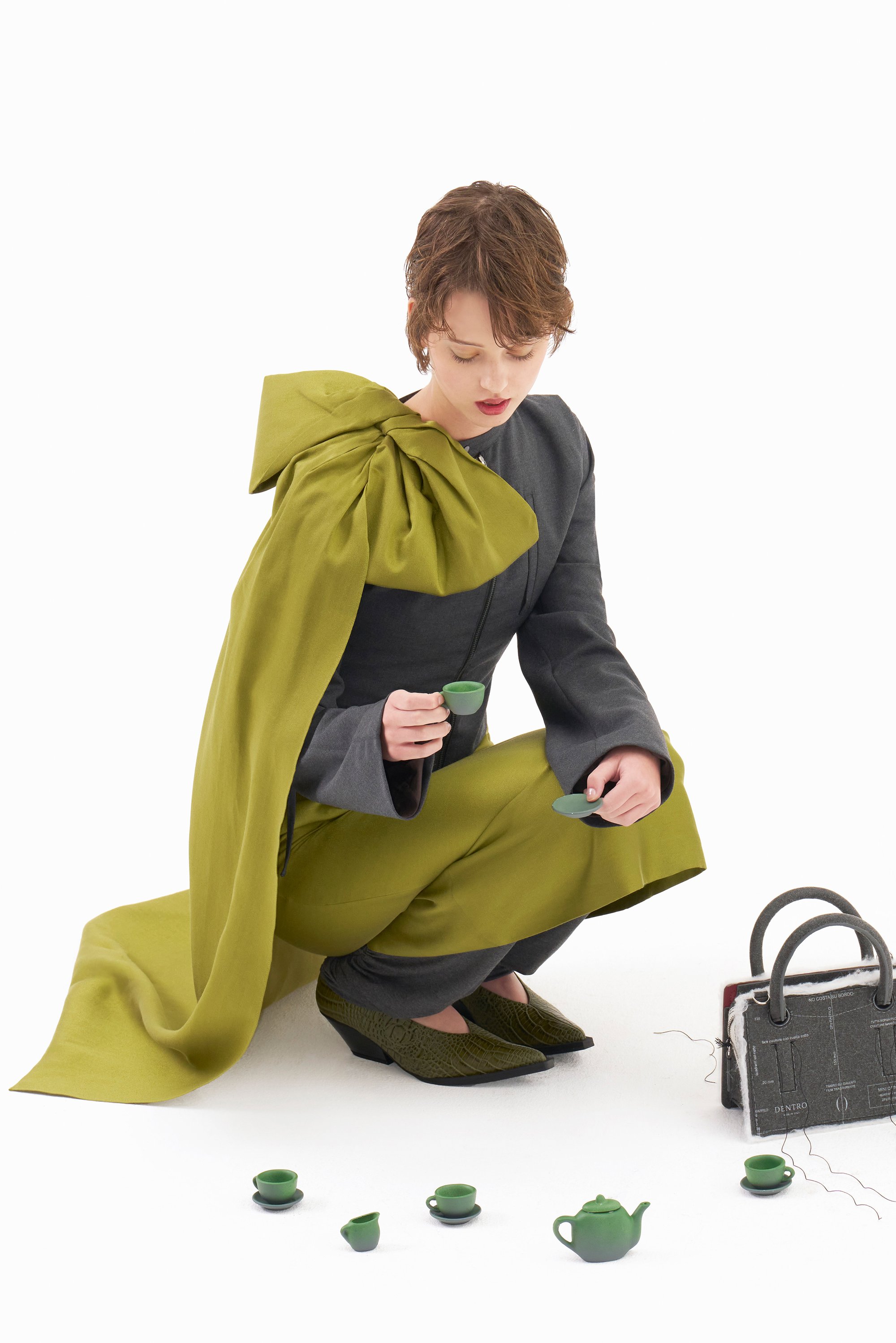 Una persona con el pelo corto, vestida con un traje gris y verde, se agacha para sostener una pequeña taza de té verde, rodeada de elementos de un juego de té en miniatura.