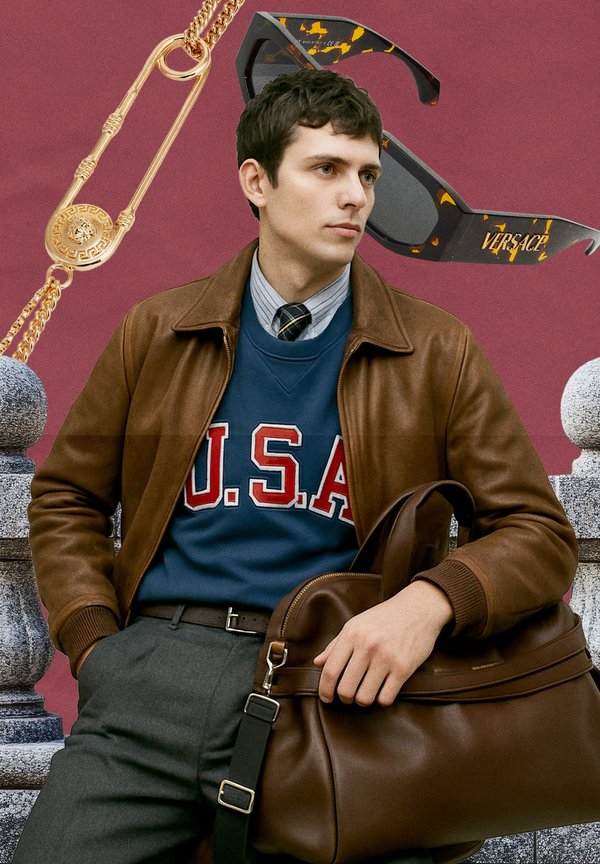 Brown leather jacket over a blue sweatshirt with "U.S.A." in red, grey trousers, and holding a brown bag; gold safety pin necklace and tortoiseshell sunglasses.
