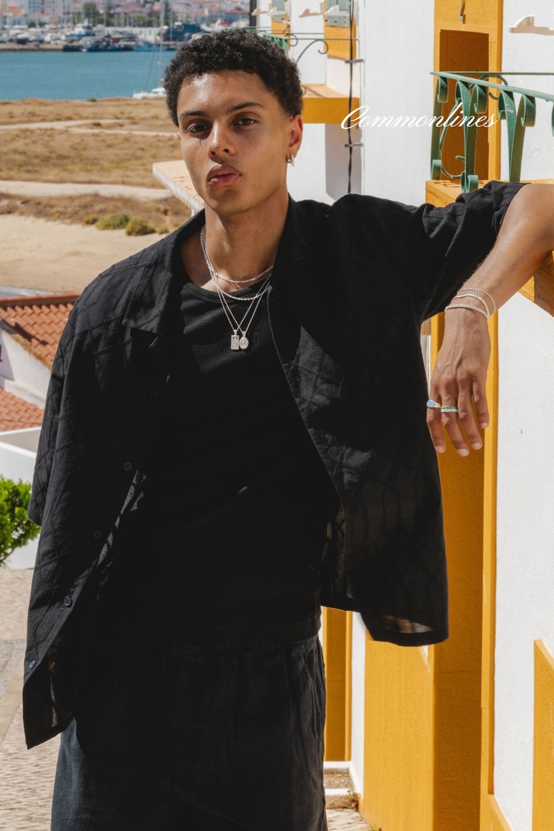 Camisa negra de manga corta con textura acolchada, superpuesta a una camiseta negra. Múltiples collares de plata adornan el cuello, realzando el aspecto.