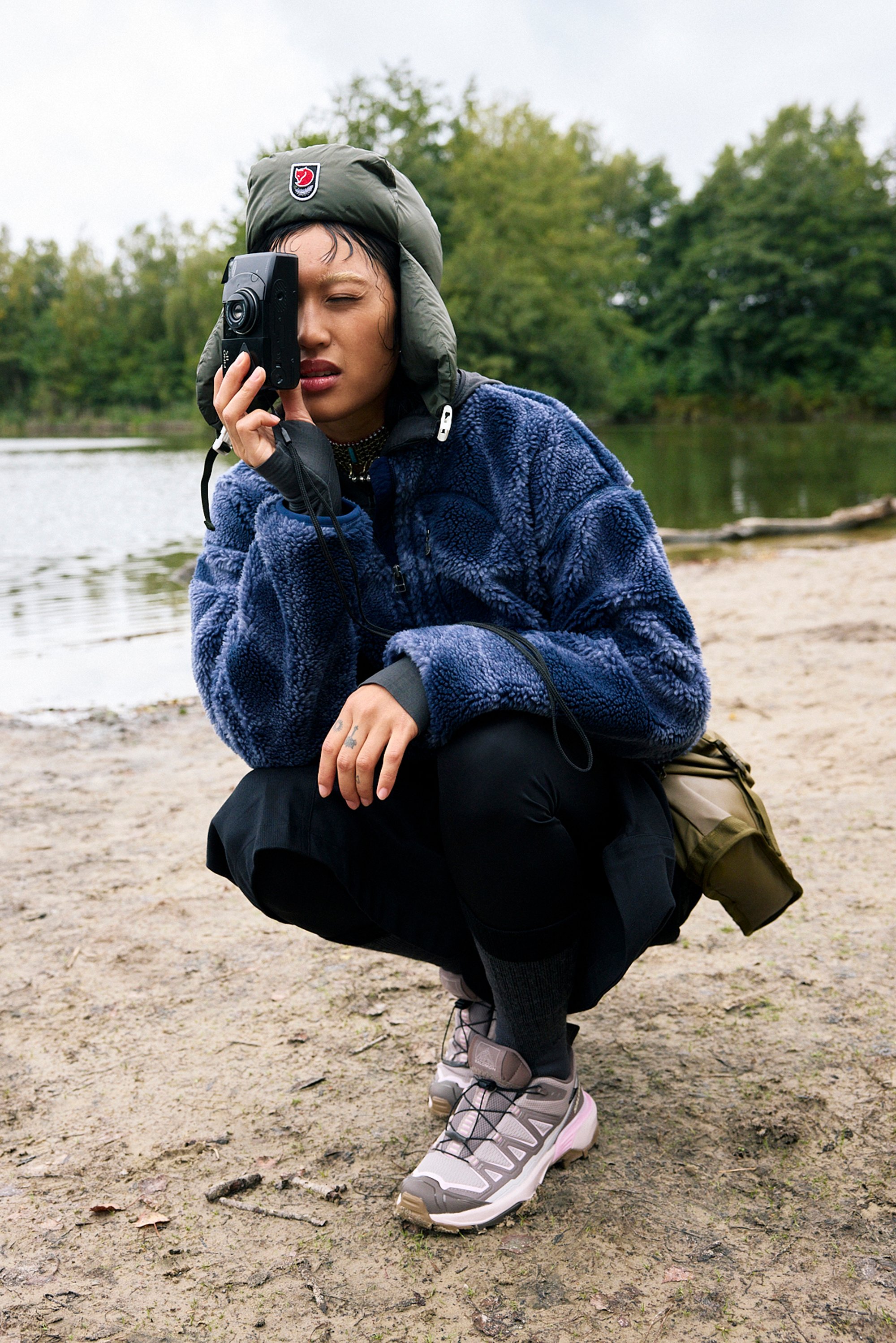 Fluffy blue jacket with a zip, black trousers, grey trainers with pink accents. Person crouching, holding a camera, by a body of water.