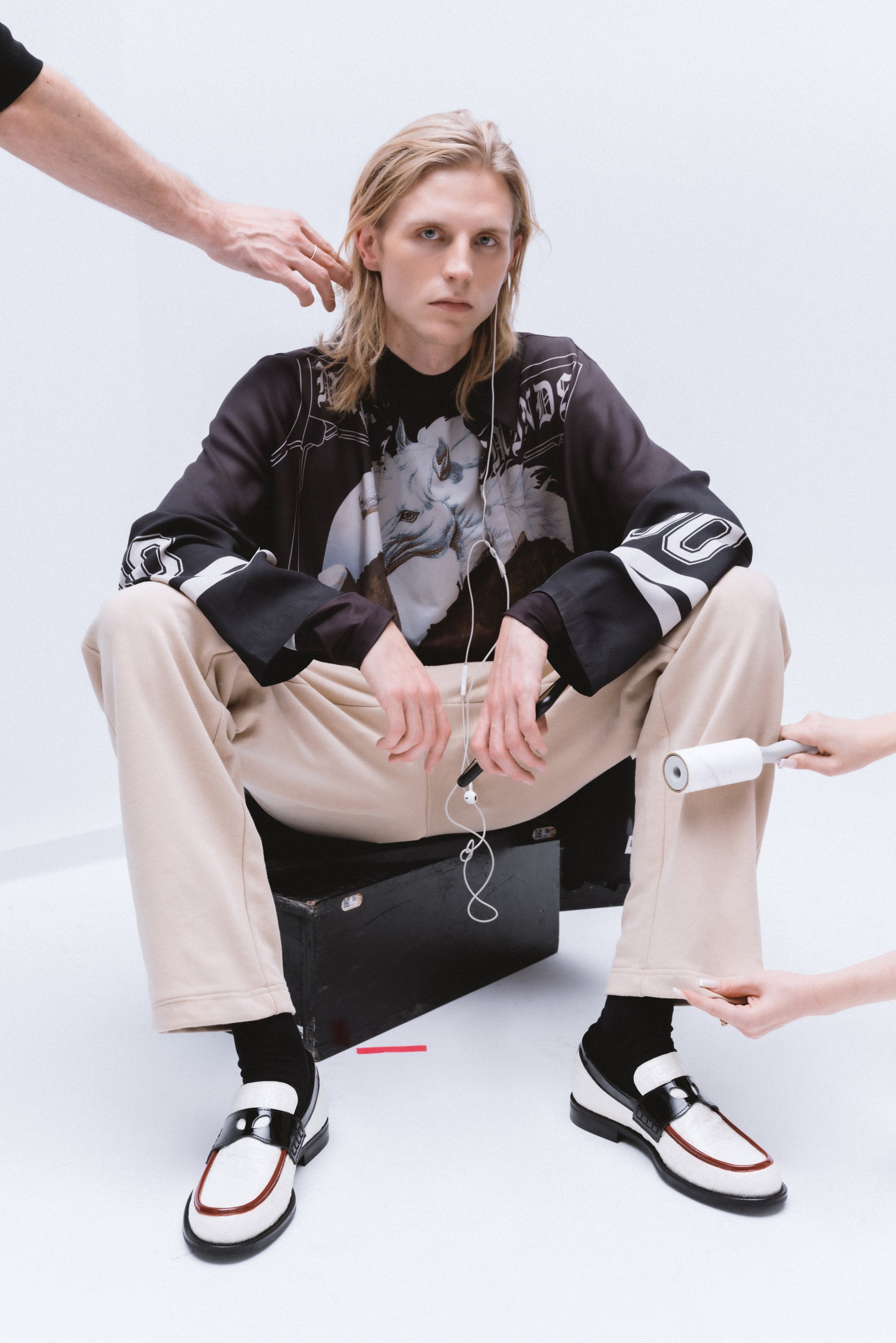 Model with long hair sits on a black box, wearing a graphic shirt and beige pants, while hands adjust his earphones and use a lint roller.