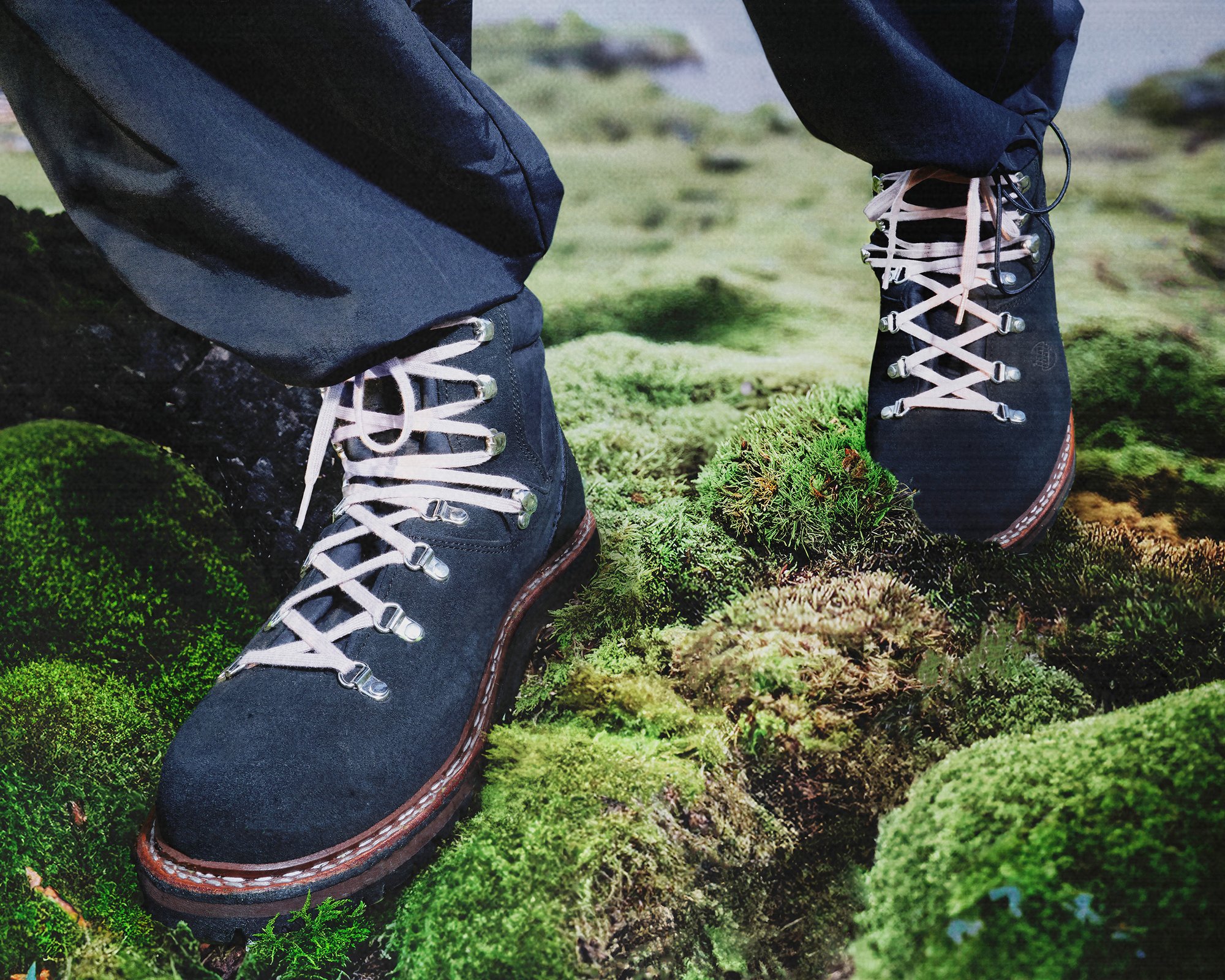 A close-up of robust black hiking boots with pink laces resting on lush green moss, indicating an outdoor trekking environment.