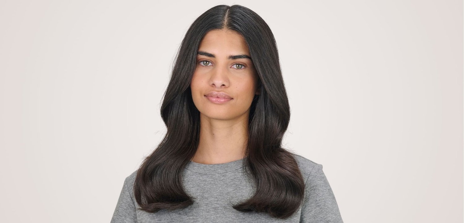 Jonge vrouw met lang donker haar, die een grijs hemd draagt, kijkt recht vooruit met een neutrale uitdrukking tegen een effen achtergrond.