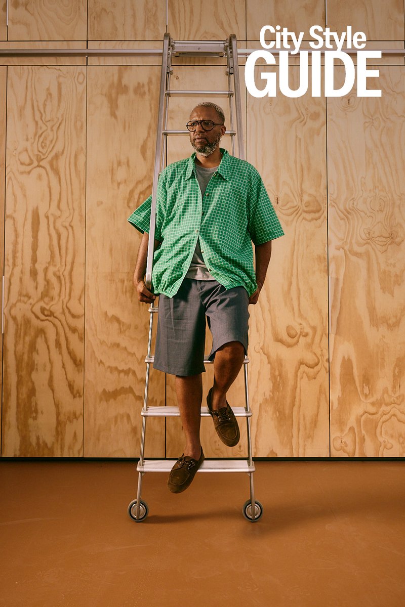 Man in green checkered shirt and shorts sitting on a rolling ladder in front of a wooden wall with "City Style Guide" text above.
