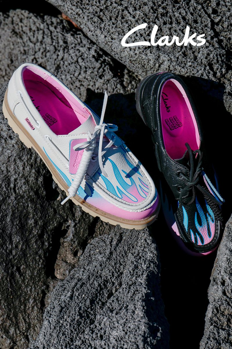 Une chaussure Clarks blanche et une noire avec des motifs de flammes bleues et des semelles roses posées sur des rochers rugueux et texturés foncés.
