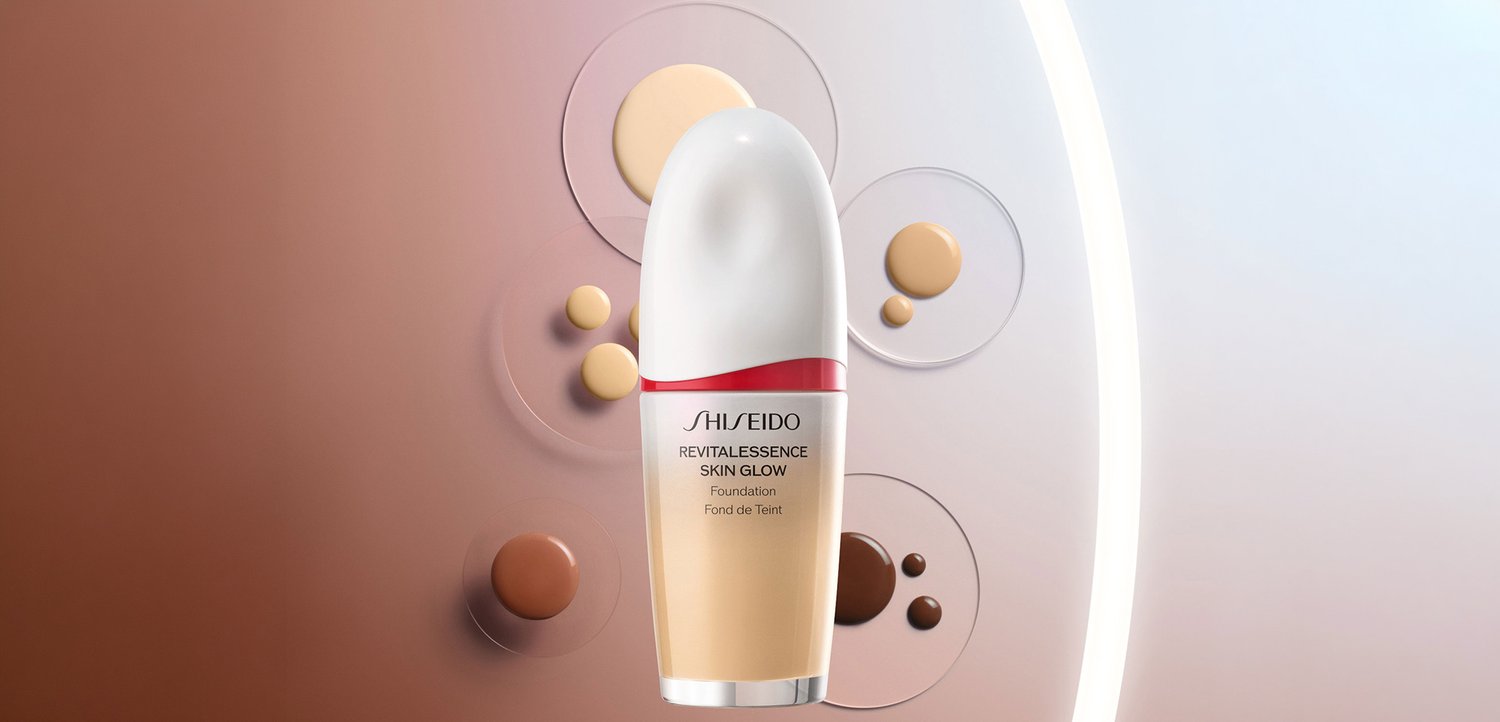 Bottiglia di fondotinta Shiseido Revitalessence Skin Glow circondata da gocce di varie tonalità di fondotinta su uno sfondo degradé.