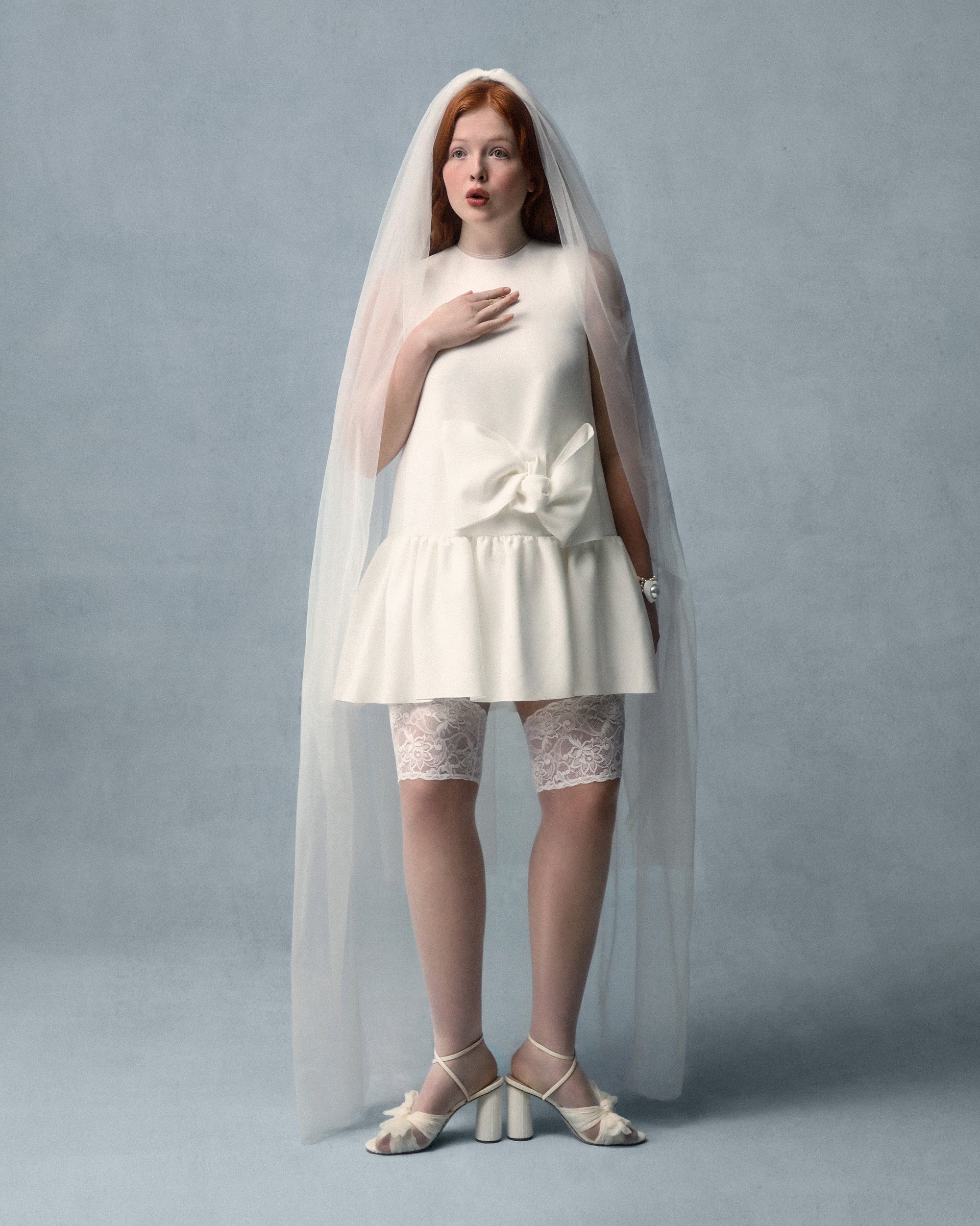Vestido mini blanco con detalle de lazo, velo transparente, medias de encaje hasta la rodilla y tacones con tiras. Tela suave y texturas suaves; diseño minimalista.