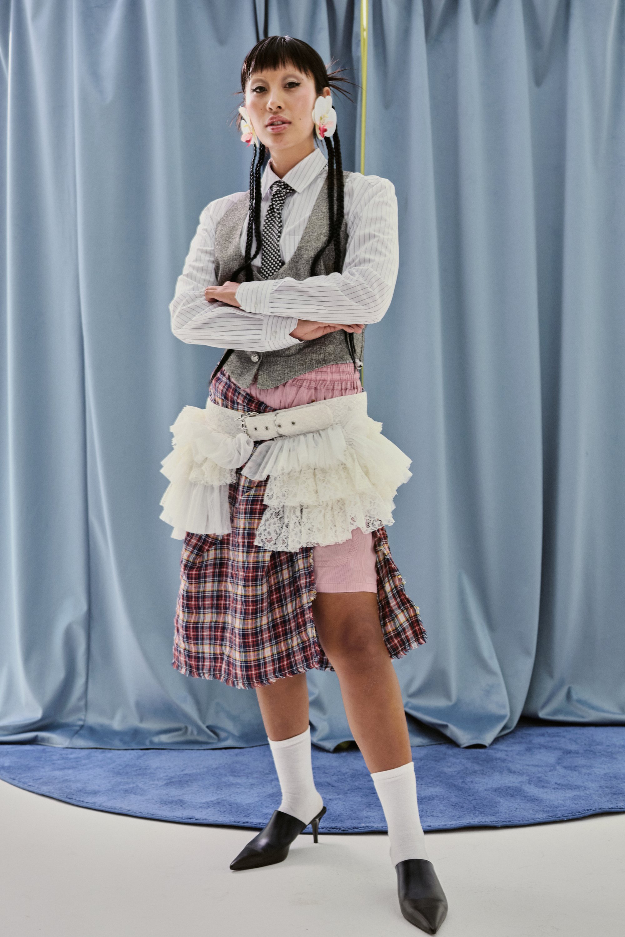 Il modello sta in piedi con sicurezza, con le braccia incrociate, indossando un outfit stratificato di plaid e pizzo, abbinato a tacchi alti, davanti a uno sfondo di tende blu.