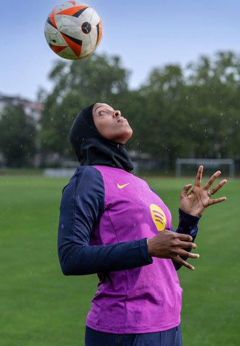 Una persona che indossa una maglia sportiva viola e blu con logo, che bilancia un pallone da calcio arancione e nero mentre sta in piedi sull'erba.