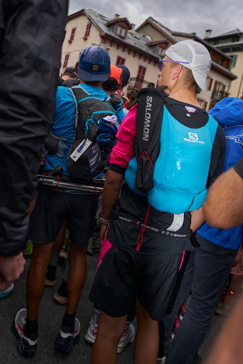Salomon hydratatietas in turquoise en zwart met verstelbare banden, gedragen over een zwart shirt, omringd door hardlopers in verschillende kleurrijke sportkleding.