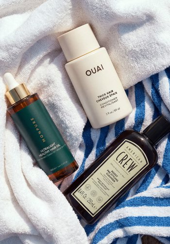 Trois flacons de soin capillaire – un flacon brun compte-gouttes, un après-shampoing épais OUAI blanc, et un nettoyant coiffant American Crew brun – sur des serviettes.