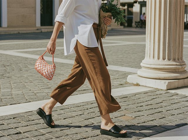 Personne marchant dans une rue pavée portant un pantalon marron, une chemise blanche, des sandales noires glissées, tenant un sac en filet et un bouquet de verdure.