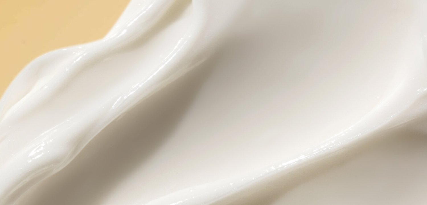 Gladde witte romige textuur met zachte ribbels, die lijkt op een huidverzorgings- of cosmetisch product dat op een oppervlak is uitgesmeerd.