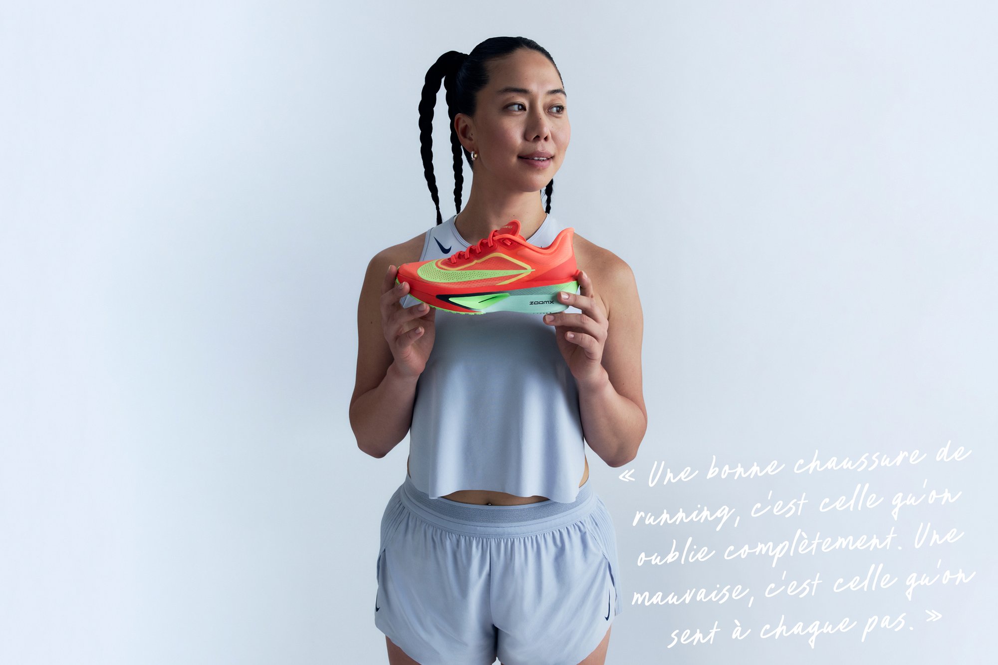 Baskets de course orange vif avec une tige en mesh, accents verts et semelle rembourrée. Modèle portant une tenue de sport grise, tenant la chaussure.