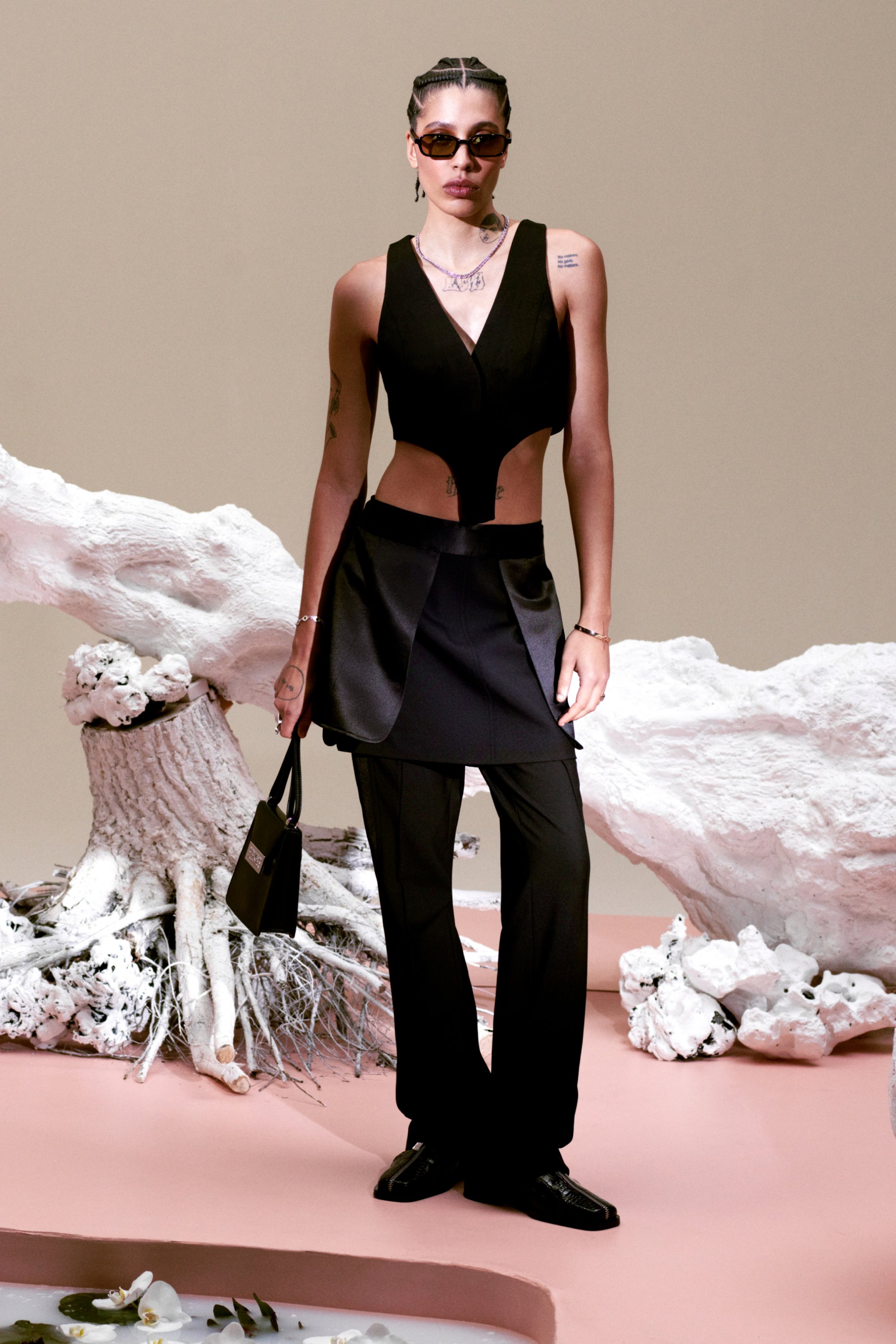A model stands confidently in a stylish black outfit with a cropped top and tailored pants, holding a small handbag, against a textured background.