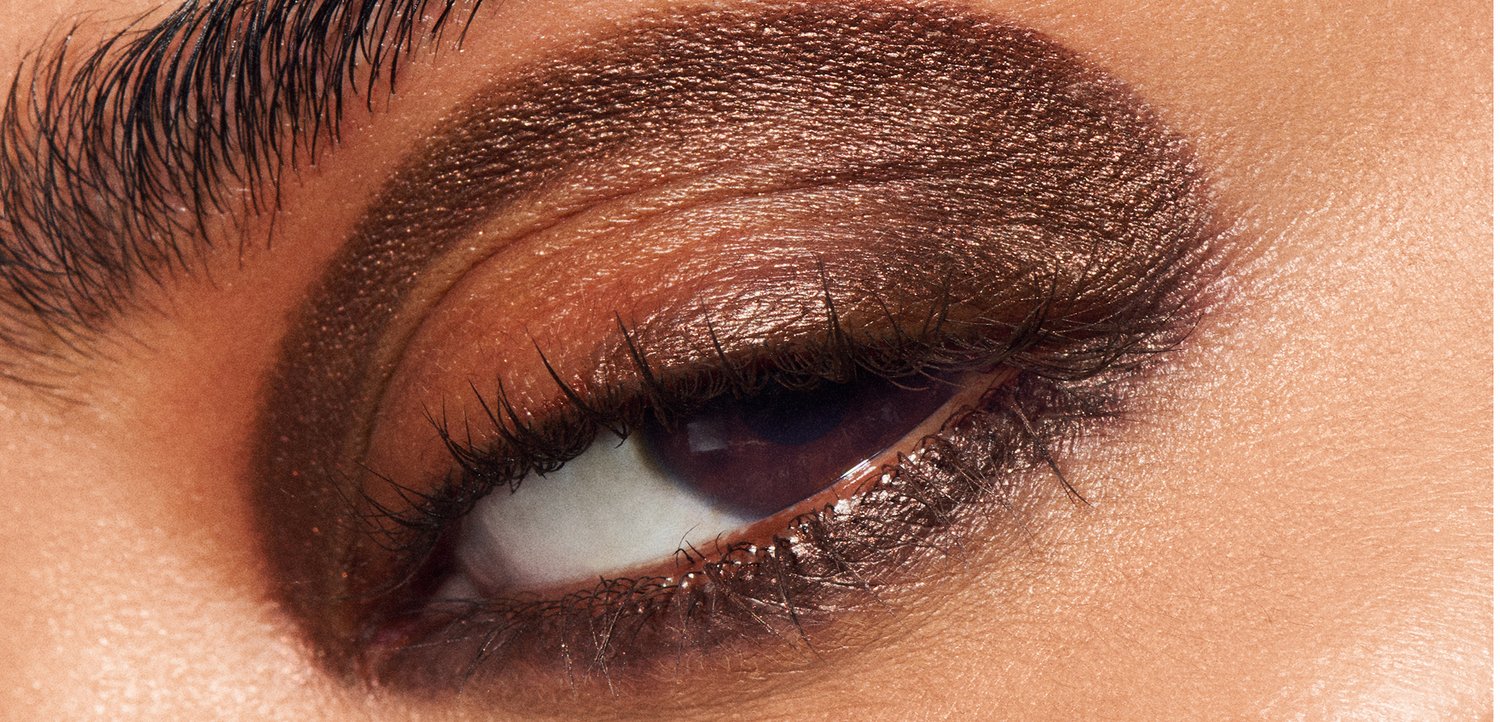Gros plan sur un œil avec un fard à paupières métallique bronze, de longs cils foncés et une texture de peau lisse avec des reflets subtils.