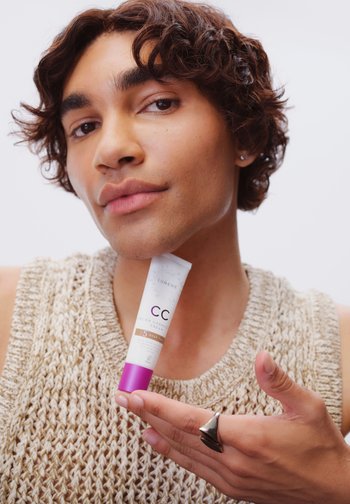 Persoon met krullend haar en gebreide mouwloze top houdt een tube Lumene CC kleurcorrigerende crème dicht bij haar kin.