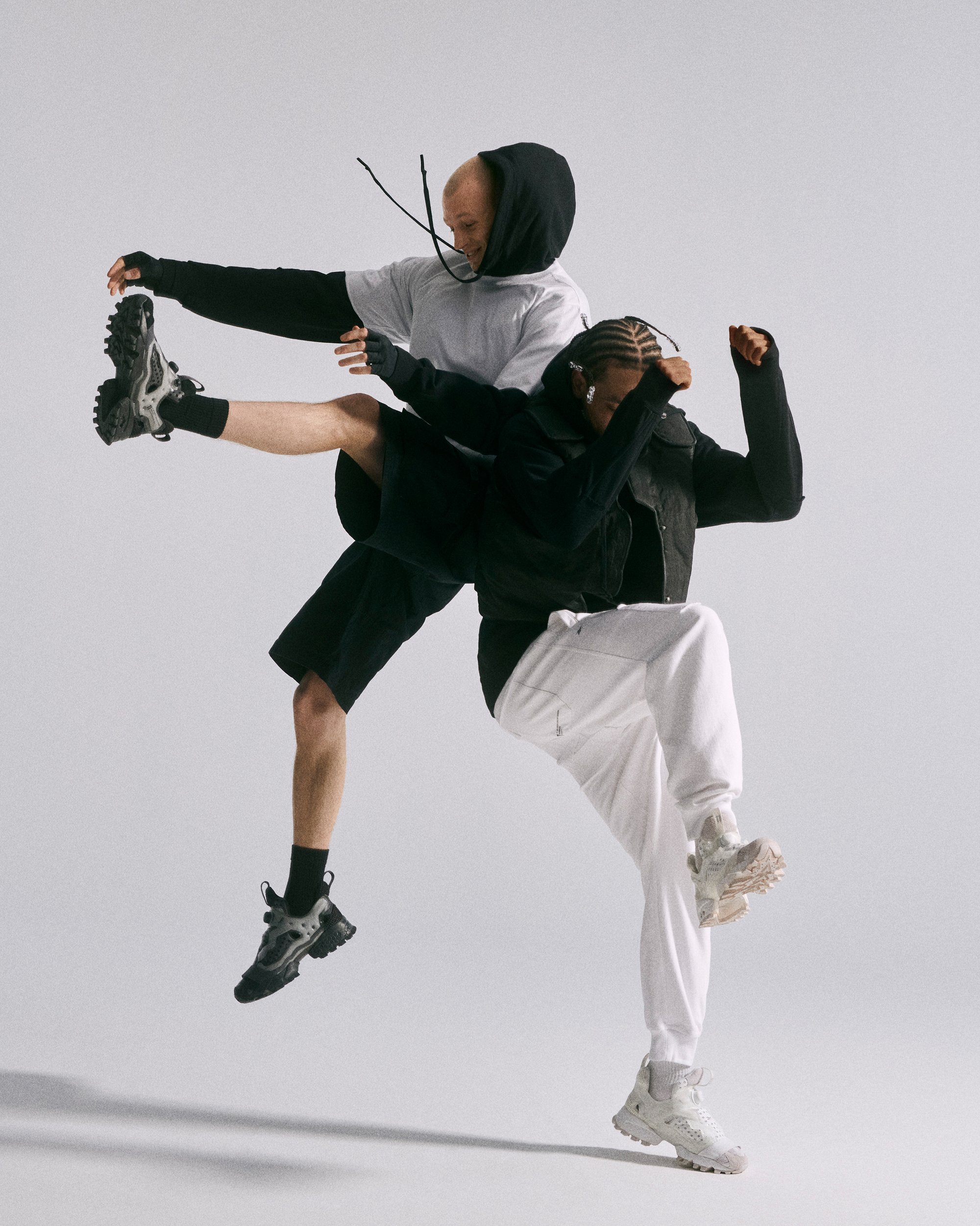 Dos bailarines saltan y patean en poses dinámicas, mostrando ropa atlética en un estudio minimalista con un fondo gris claro.