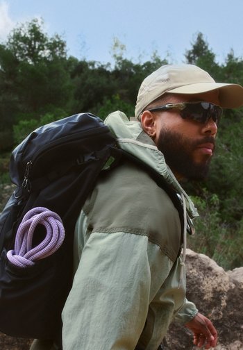 Man wearing sunglasses, cap, and green jacket hiking with a black backpack and coiled rope, forest in the background.