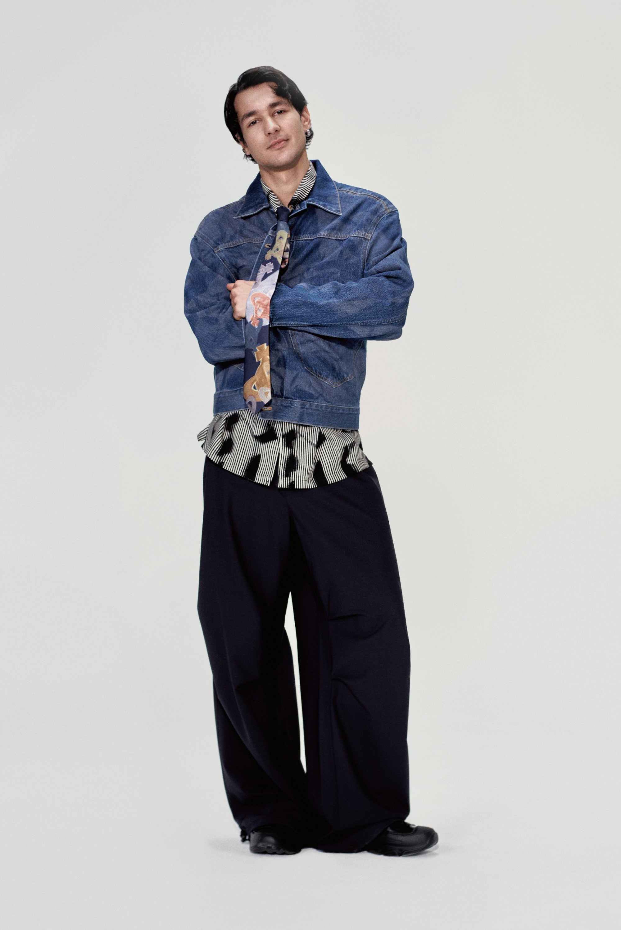 A young man stands confidently, wearing a denim jacket over a patterned shirt and wide black trousers, arms crossed and smiling.