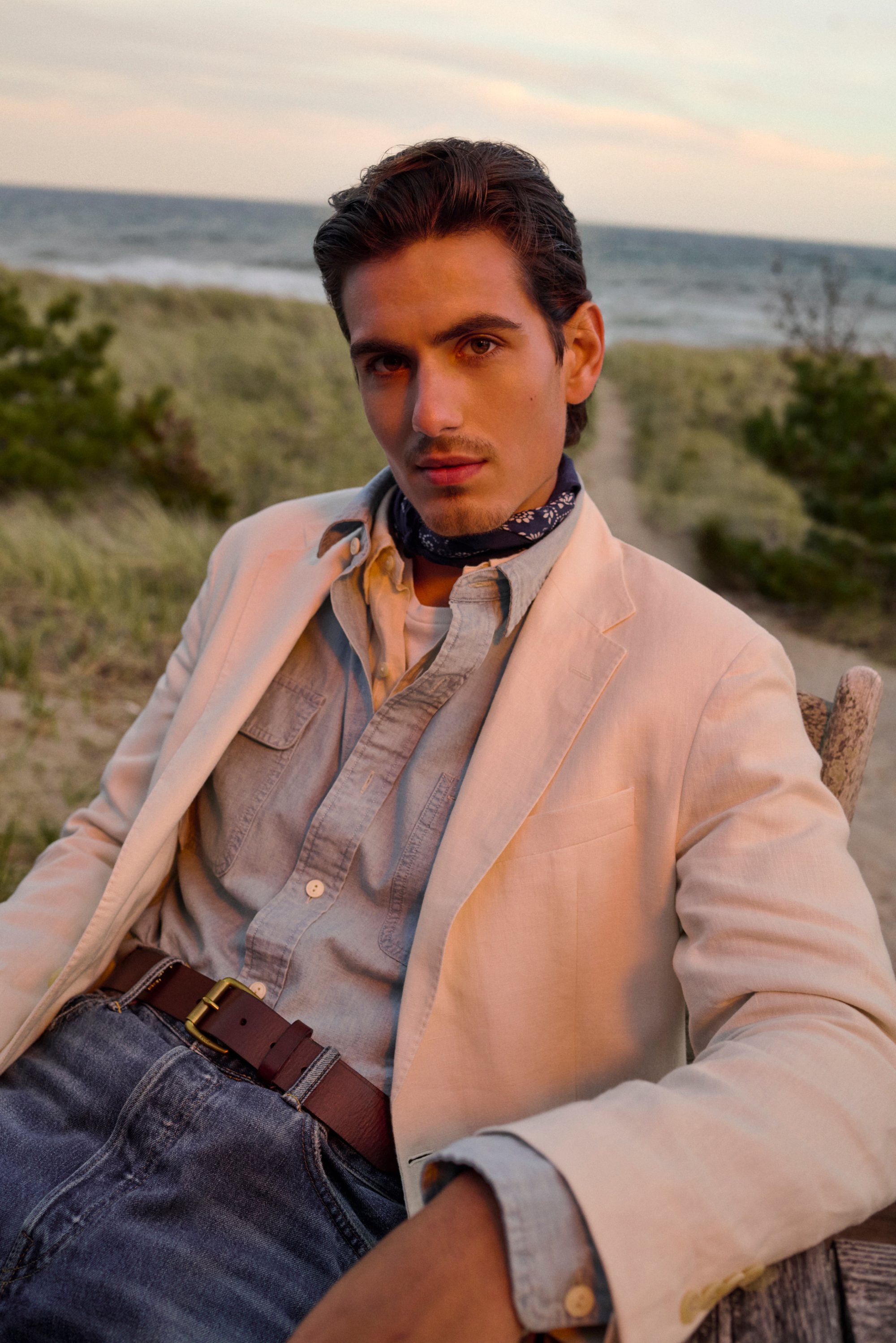 Un joven con el cabello estilizado se sienta en una silla de madera junto a hierba de playa, vistiendo una chaqueta ligera, una camisa de mezclilla y una bufanda, con el océano de fondo.
