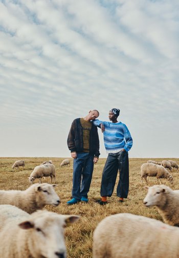 Twee personen staan in een grasveld met schapen. De een draagt een donkere cardigan over een gele trui; de ander heeft een blauw gestreepte trui en losse broek aan.