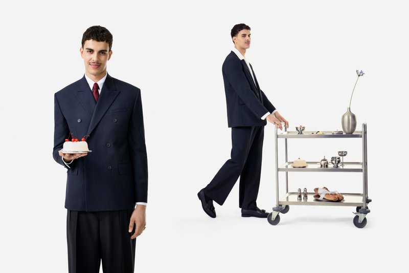 Ein Mann im dunklen Anzug hält einen kleinen Kuchen mit Kirschen und schiebt einen Metallwagen mit einer Vase, Geschirr, Brot und einem weiteren Kuchen.