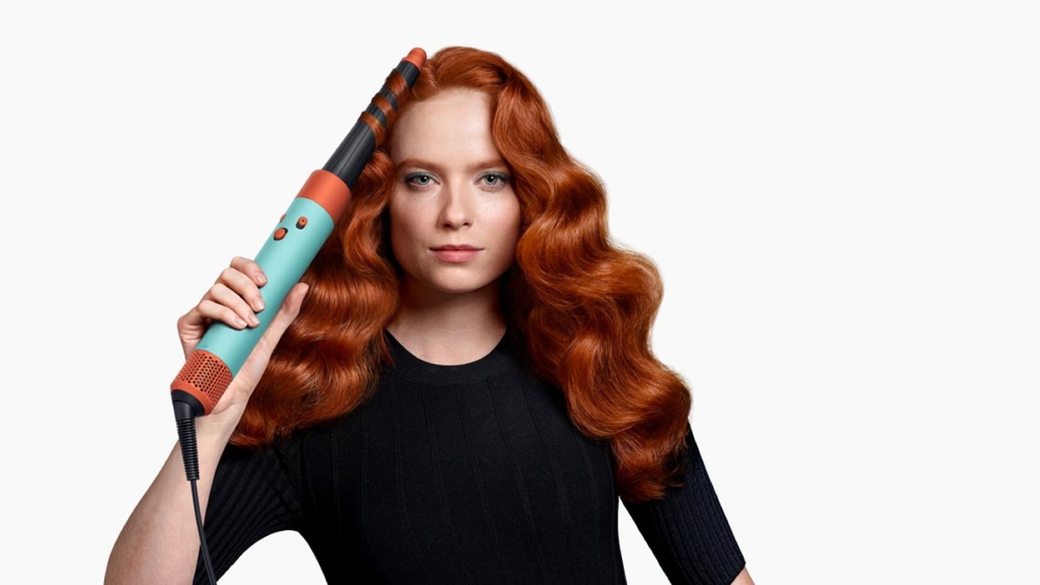 Donna con lunghi capelli rossi ondulati tiene in mano un ferro arricciacapelli turchese e arancione mentre acconcia una ciocca di capelli su uno sfondo bianco uniforme.