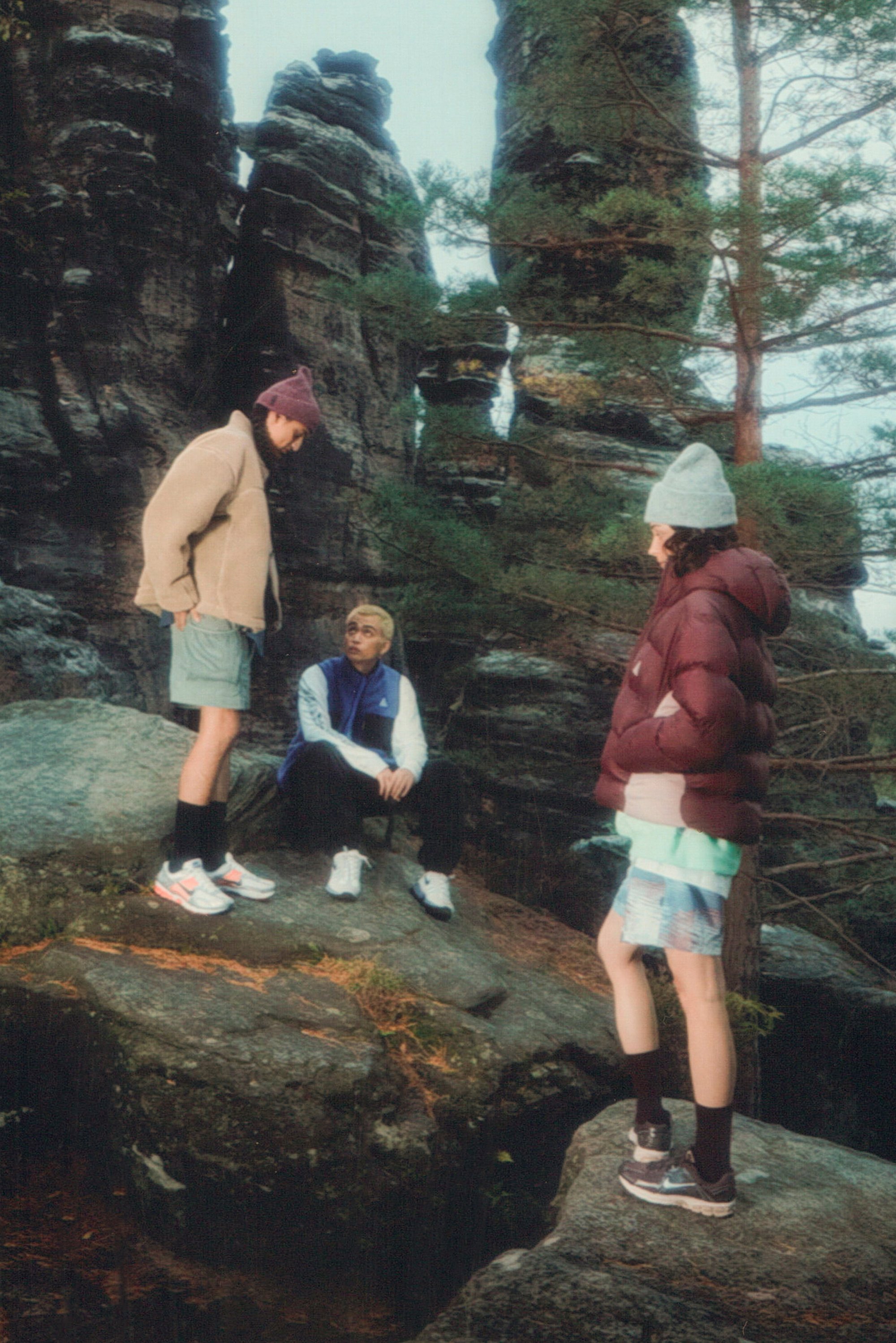 Three young people in casual outdoor attire gather on large rocks surrounded by tall trees and stone formations, engaged in conversation.