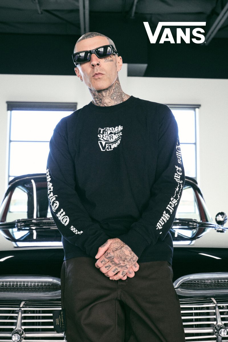 Hombre con tatuajes y gafas de sol está delante de un coche vintage negro, vistiendo una camiseta de manga larga negra de Vans con letras blancas en las mangas y el pecho.
