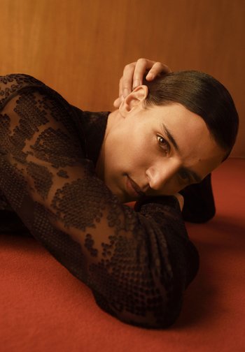 Person with slicked-back hair and sheer black patterned top rests head on crossed arms against a reddish surface and wooden background.