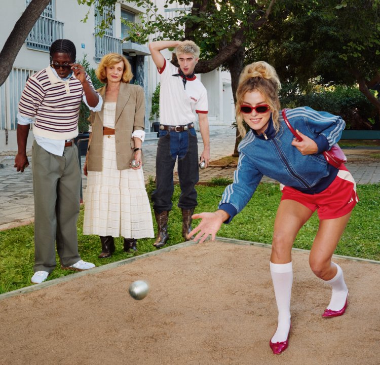 Quatre personnes en plein air ; une femme en short rouge lance une boule métallique tandis que trois autres regardent sur un terrain en terre près de la verdure et des bâtiments.