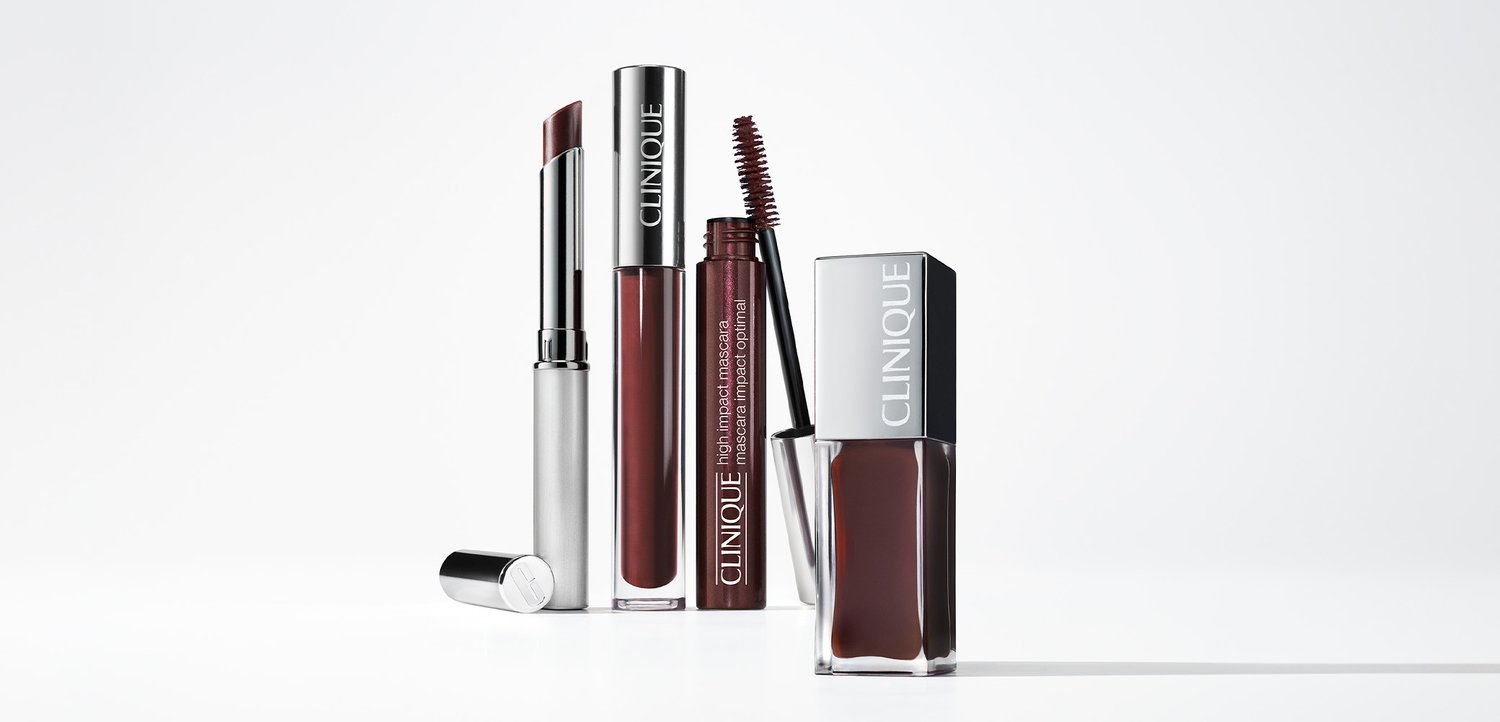 Vier Clinique-Kosmetikprodukte auf weißem Hintergrund: ein silberner Lippenstift, ein dunkelroter flüssiger Lippenstift, eine Wimperntusche in einer eleganten Flasche und eine quadratische Nagellackflasche.