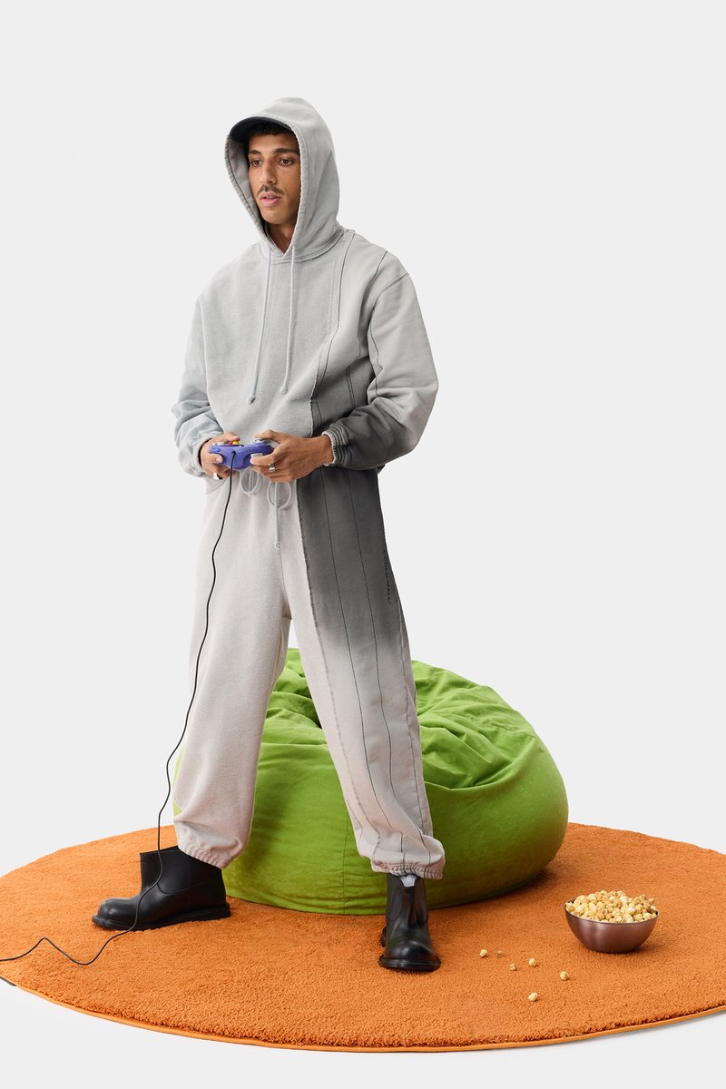 Monopieza de sudadera gris con capucha, con rayas oscuras. Botas oscuras, alfombra redonda naranja, puf verde y un tazón de palomitas cerca.