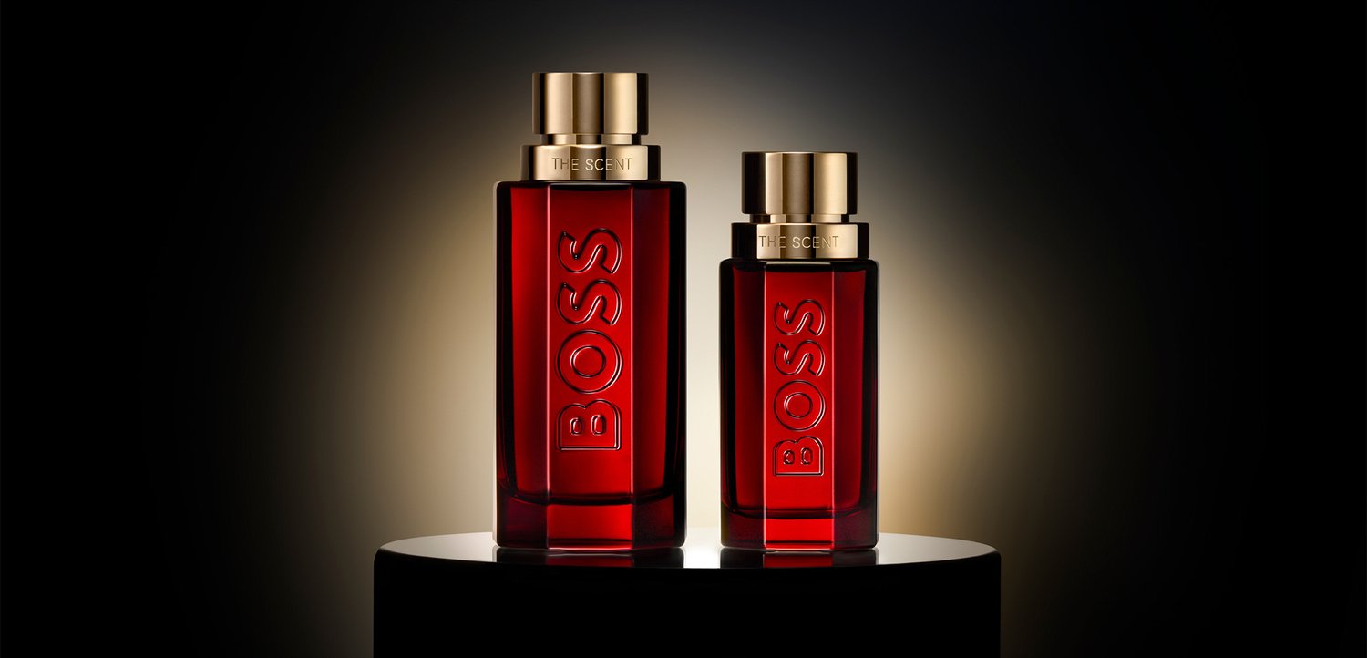 Due bottiglie di profumo rosse rettangolari con tappi dorati etichettate BOSS THE SCENT, esposte su un piedistallo tondo nero con sfondo scuro.