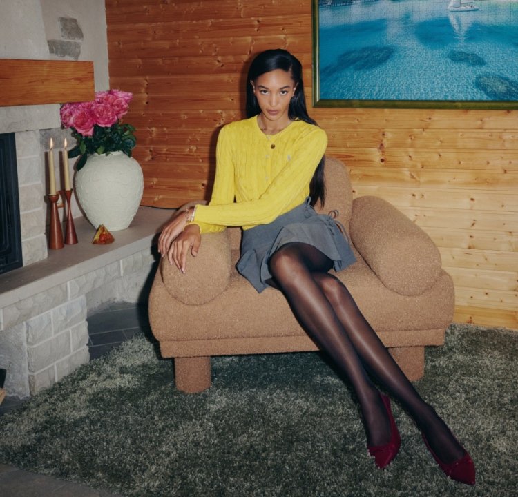 Brun soffa med korkstruktur och rundade armstöd, modell i gul stickad tröja och grå kjol, svarta strumpbyxor och vinröda högklackade skor, blomsterdekoration.