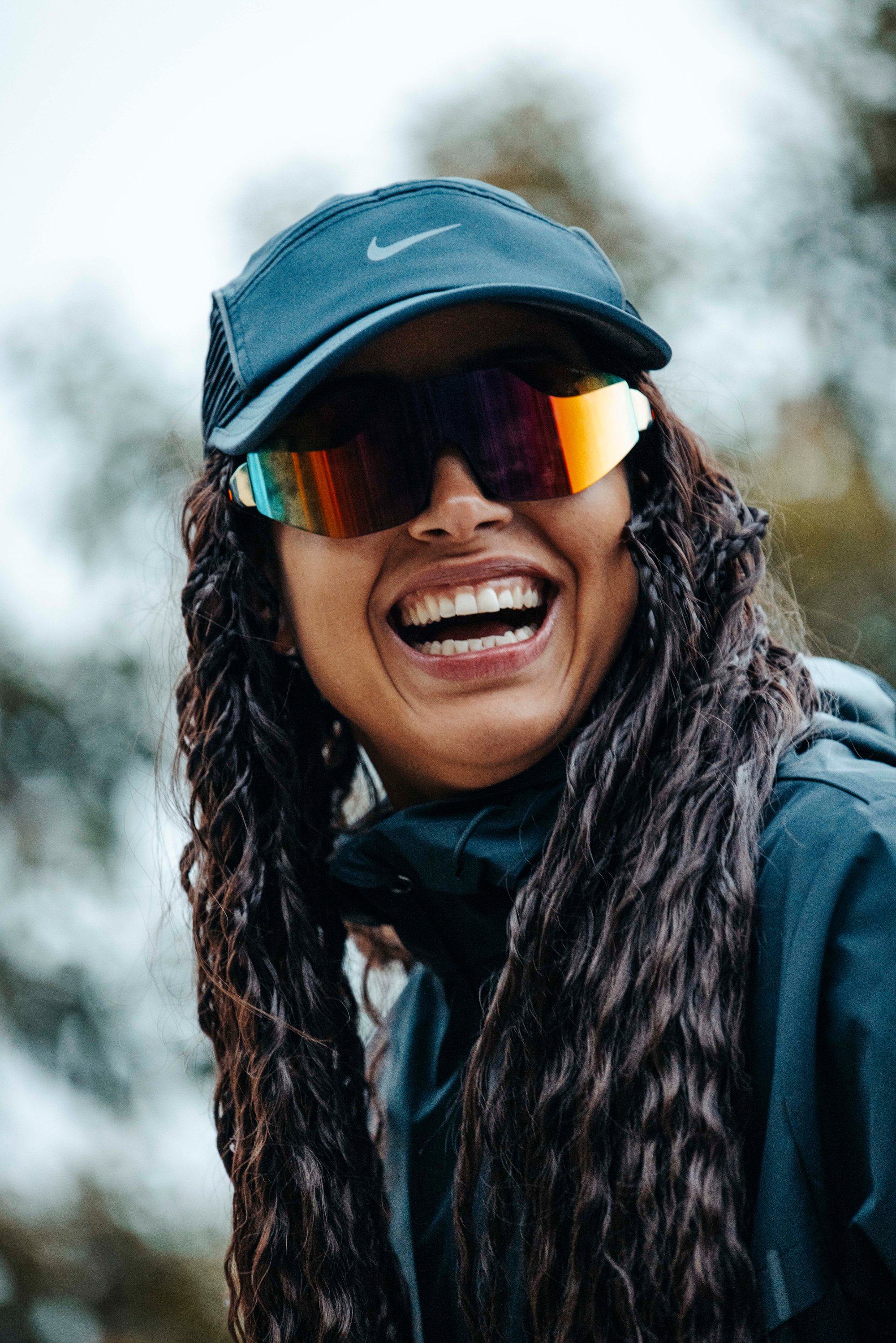 Persona sonriente con cabello largo y rizado que lleva una gorra de Nike y gafas de sol coloridas, contra un fondo exterior desenfocado.
