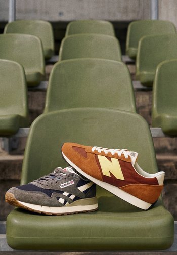 Two sneakers, one grey Reebok and one orange New Balance, rest on a green stadium seat with empty rows in the background.